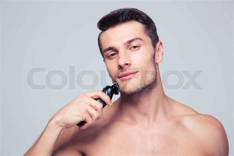 Handsome young man shaving with electric razor over gray background and looking at camera, stock photo