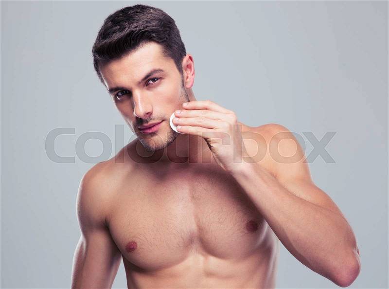 Handsome man cleaning face skin with batting cotton pads over gray background and looking at camera, stock photo