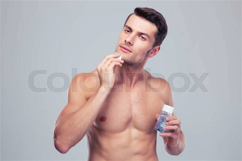 Handsome man applying lotion after shave on face over gray background, stock photo