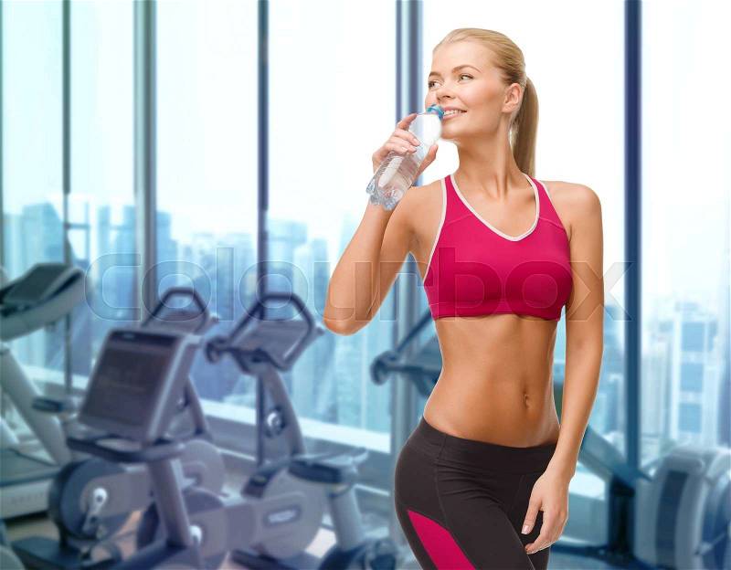 People, sport, fitness and recreation concept - happy woman drinking water from bottle over gym machines background, stock photo