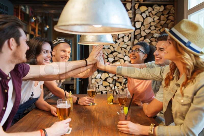 People, leisure, friendship and communication concept - group of happy smiling friends with drinks making high five gesture at bar or pub, stock photo