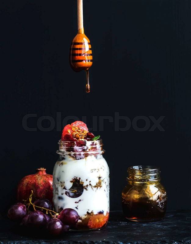 Yogurt and oat granola with grapes, pomegranate and grapefruit in a tall glass jar on black backdrop. Hand pours honey over the jar with a wooden stick, stock photo