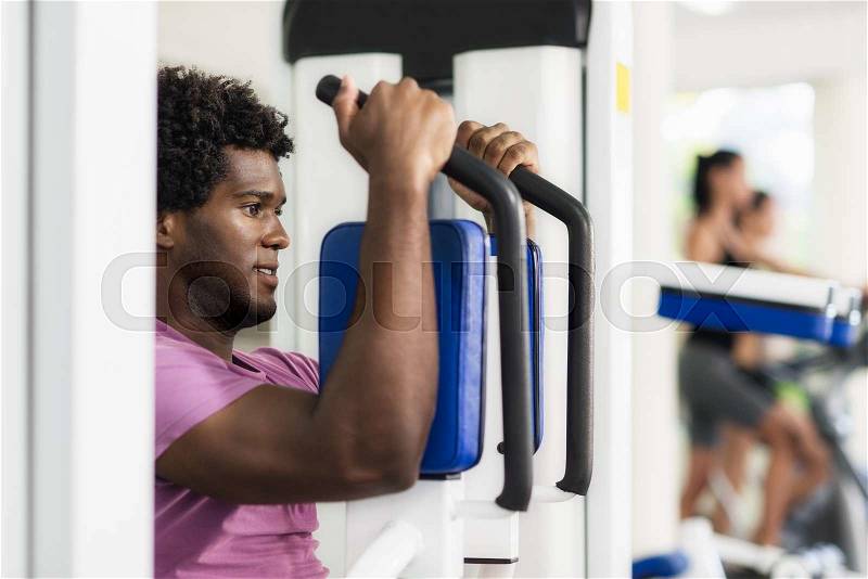 Young black man exercising pectoral muscles in fitness club, with people working out on cyclette in background, stock photo