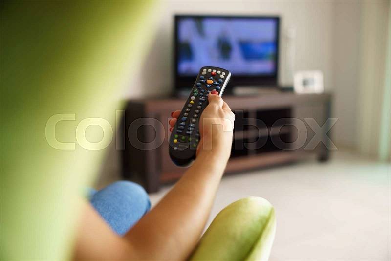 Over the shoulder view of girl sitting on sofa holding tv remote and surfing programs on television, stock photo