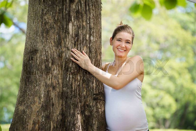 Ecology and environment-Portrait of white pregnant woman in t-shirt embracing and hugging tree in park, smiling and looking at camera, stock photo