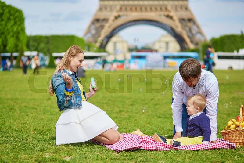 Happy family of three, mother, father and little toddler boy, having picnic in Paris near the Eiffel tower and playing with bubbles, stock photo