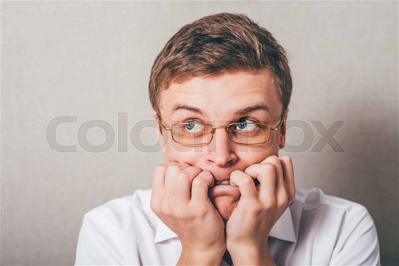 The man with glasses biting his fists, bites his nails. On a gray background, stock photo