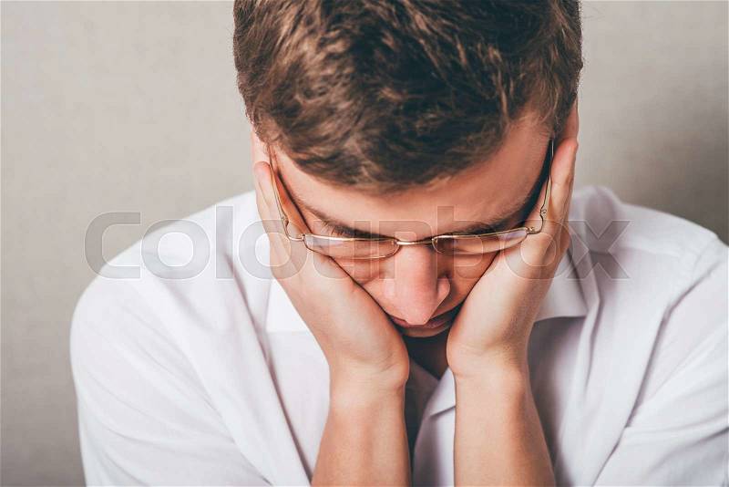 The man in glasses upset sad, hands on his cheeks, his head lowered. On a gray background, stock photo