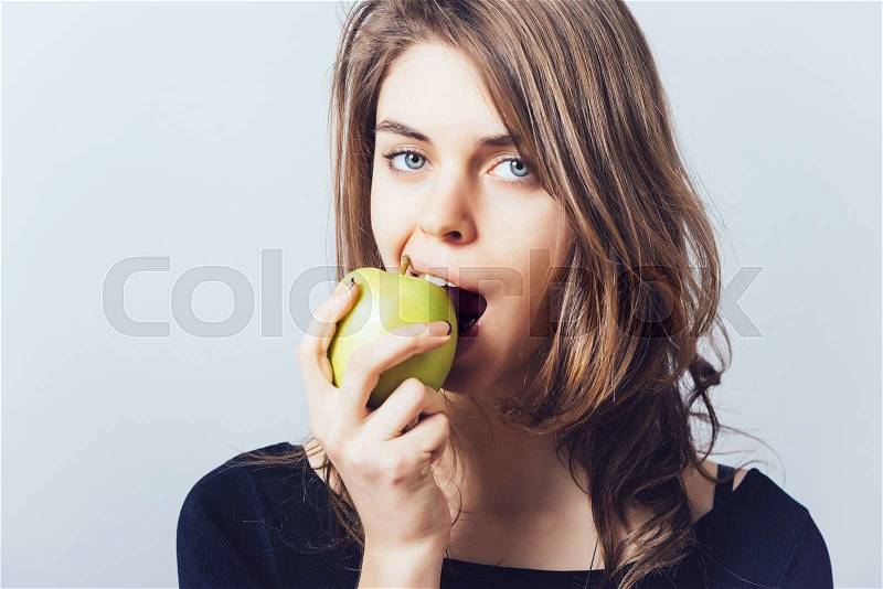 Beautiful young woman bites a green apple on a gray background, stock photo