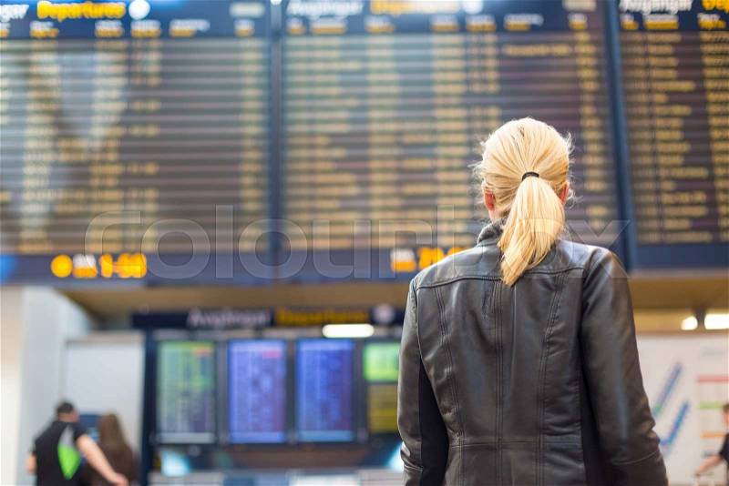 Casually dressed young stylish female traveller checking a departures board at the airport terminal hall in front of check in couters. Flight schedule display blured in the background. Focus on woman, stock photo