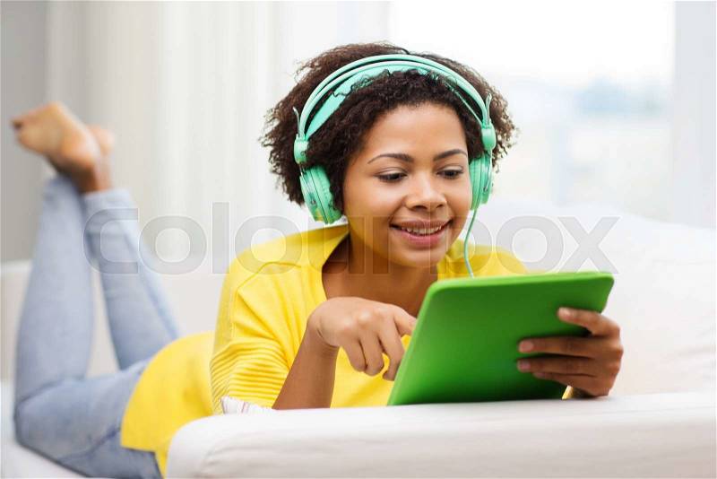 People, technology and leisure concept - happy african american young woman lying on sofa with tablet pc computer and headphones listening to music at home, stock photo