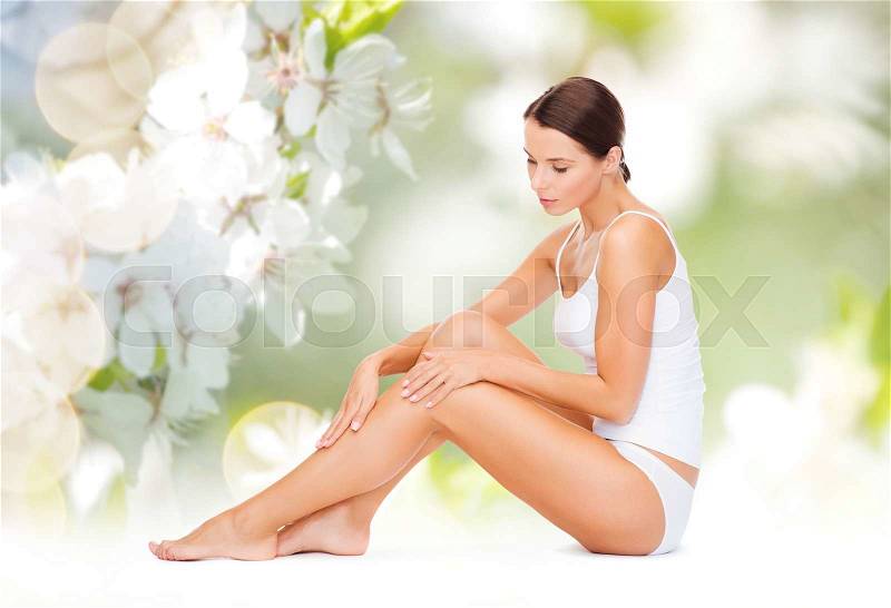People, beauty and body care concept - beautiful woman in cotton underwear touching legs over green natural cherry blossom background, stock photo