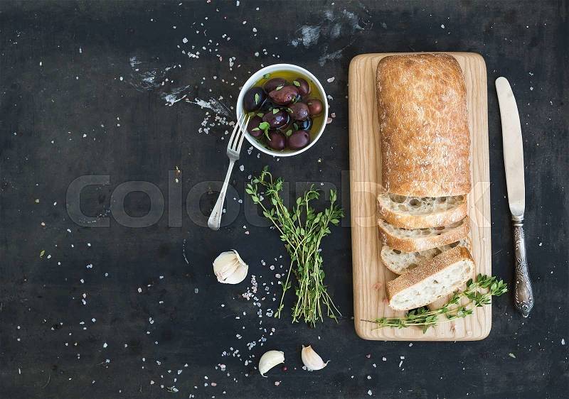 Italian ciabatta bread cut in slices on wooden chopping board with herbs, garlic and olives over dark grunge backdrop, copy space, top view, stock photo