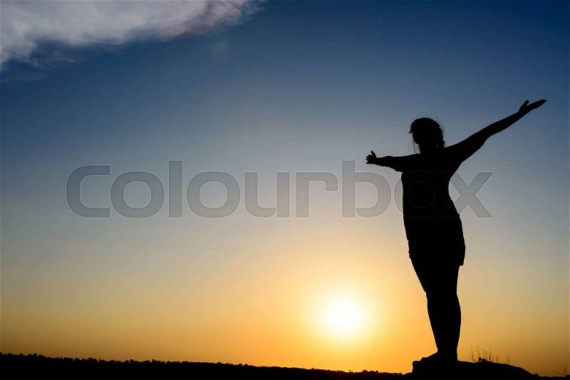 Woman embracing the sunrise or sunset standing silhouetted with outspread arms backlit by the fiery orb of the sun, spiritual image with copyspace, stock photo