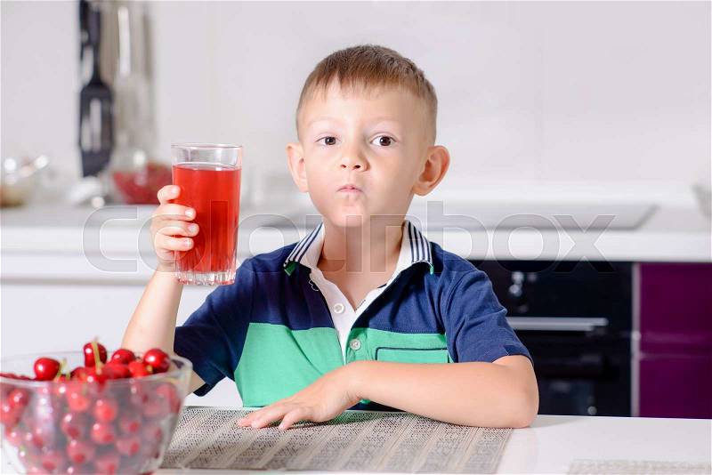 Young Blond Boy Sitting at Kitchen Table with Bowl of Ripe Cherries Drinking Tall Glass of Red Juice, stock photo