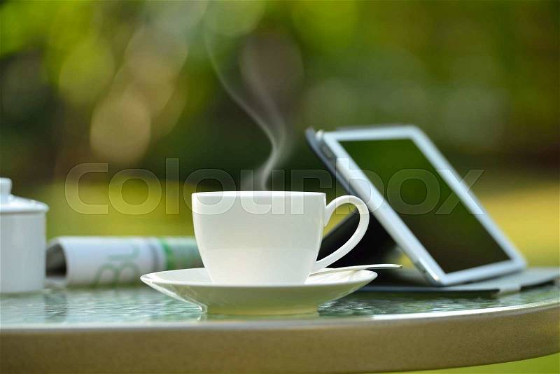 Cup of coffee with smoke and tablet computer in garden, stock photo