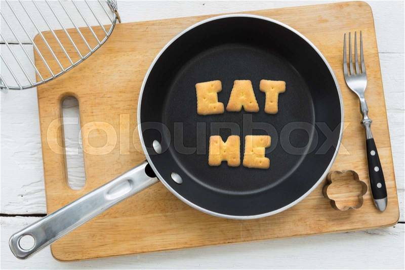 Top view of letter collage made of biscuits. Word EAT ME putting in black frying pan. Other cooking equipments: fork, cookie cutter and chopping board putting on white wooden table, vintage style image, stock photo