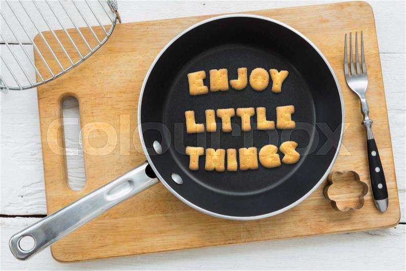 Top view of alphabet collage made of crackers. Quote ENJOY LITTLE THINGS putting in black pan. Other kitchenware: fork, cookie cutter and chopping board putting on white wooden table, vintage style image, stock photo