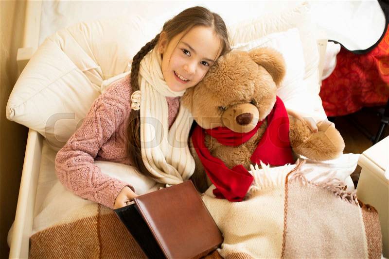 Portrait of cute smiling girl reading book with teddy bear in bed, stock photo