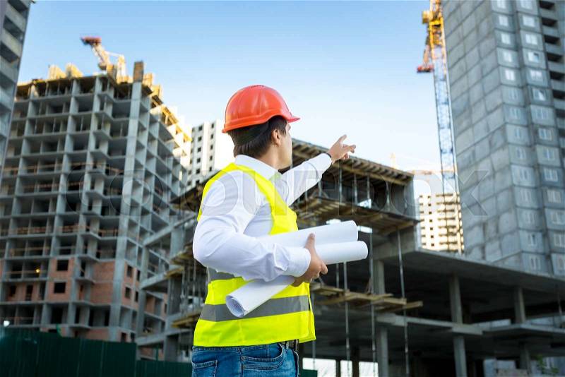Site manager in hardhat pointing at crane on building site, stock photo