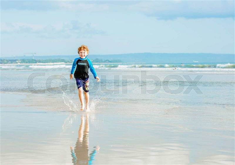 Young boy in swimming shorts and rash vest runs along Bali beach near sunset with reflection in the water, stock photo