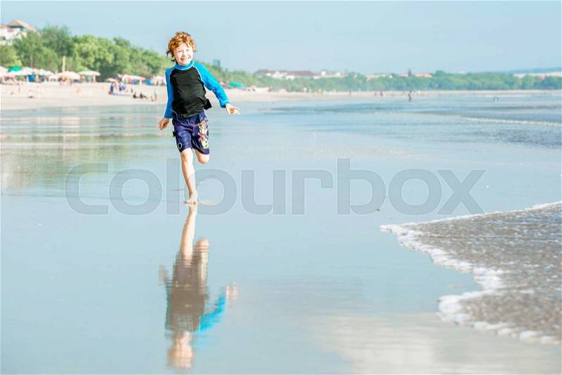Young boy in swimming shorts and rash vest runs along Bali beach near sunset with reflection in the water, stock photo