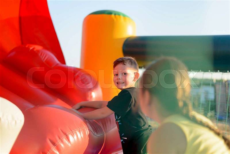 Small blonde boy (6-8 years) wearing t-shirt playing on red bouncy castle looking at camera, rear view of brunette girl (9-10 years) in foreground, stock photo