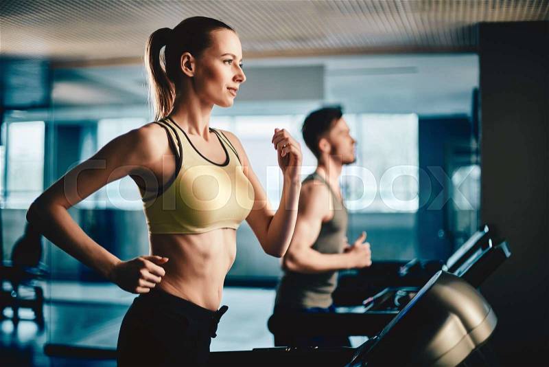 Pretty woman running on treadmill with fit young man on background, stock photo