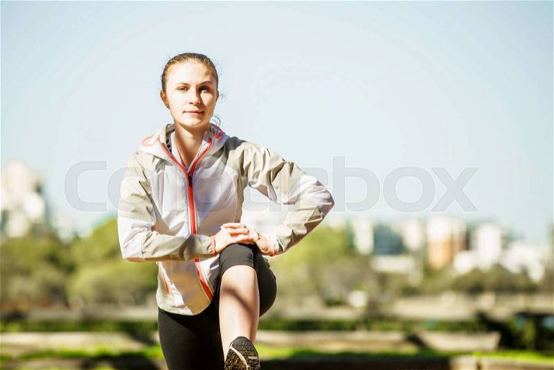 Young fit sports girl stretching out on bench before exercising in autumn pak with city at background, stock photo