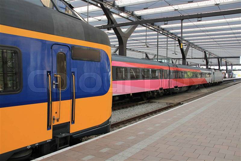 At the foreground the train of the Dutch railways and at the background the train in the colours red, pink and white at the train station, stock photo