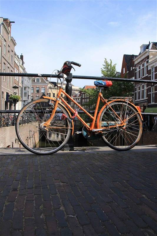 The striking locked orange painted bike stands against the fence at the canal in the city in the summer, stock photo