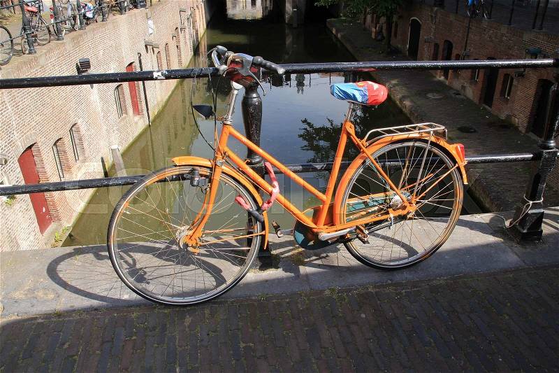The striking locked orange painted bike stands against the fence at the canal in the city in the summer, stock photo