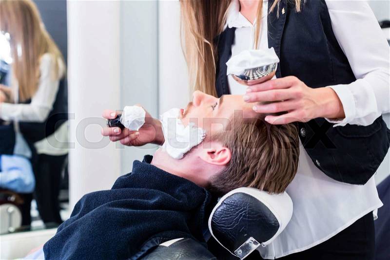 Woman barber shaving customer in her shop, applying soap with a brush, stock photo