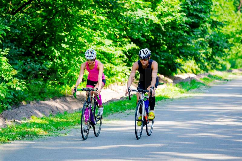 Beautiful young pair of professional cyclists in gear and glasses riding on the road surrounded by green trees on bright sunny day. Girl in pink jumpsuit with helmet, guy in dark blue special suit, stock photo