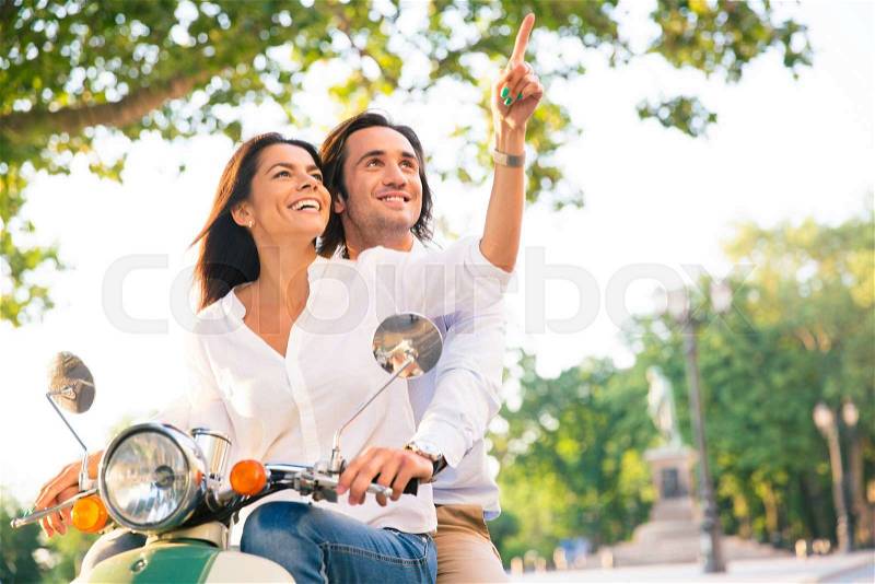 Smiling young couple on scooter together while woman pointing finger away in the town, stock photo