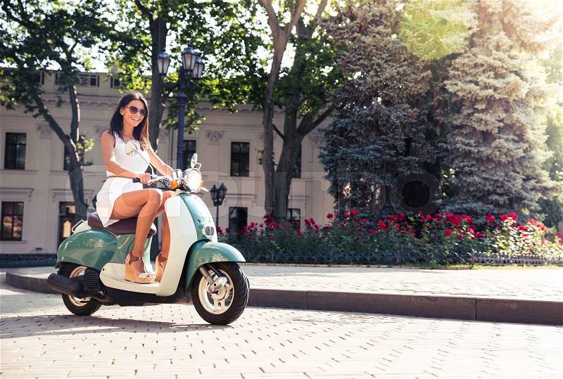 Cheerful young woman driving scooter in town, stock photo