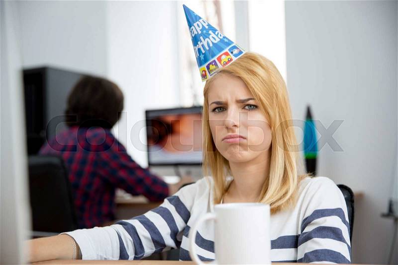 Sad woman with birthday hat sitting at the table in office and looking at camera, stock photo