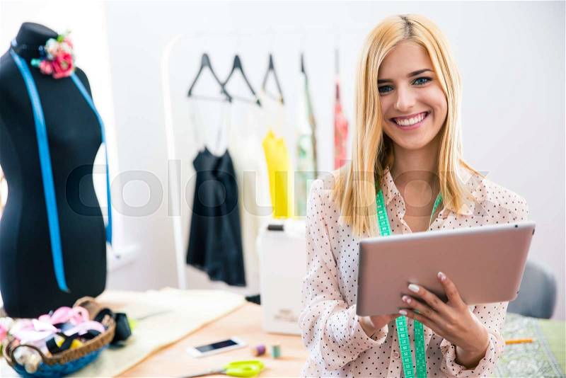 Happy female tailor holding tablet computer in workshop and looking at camera, stock photo