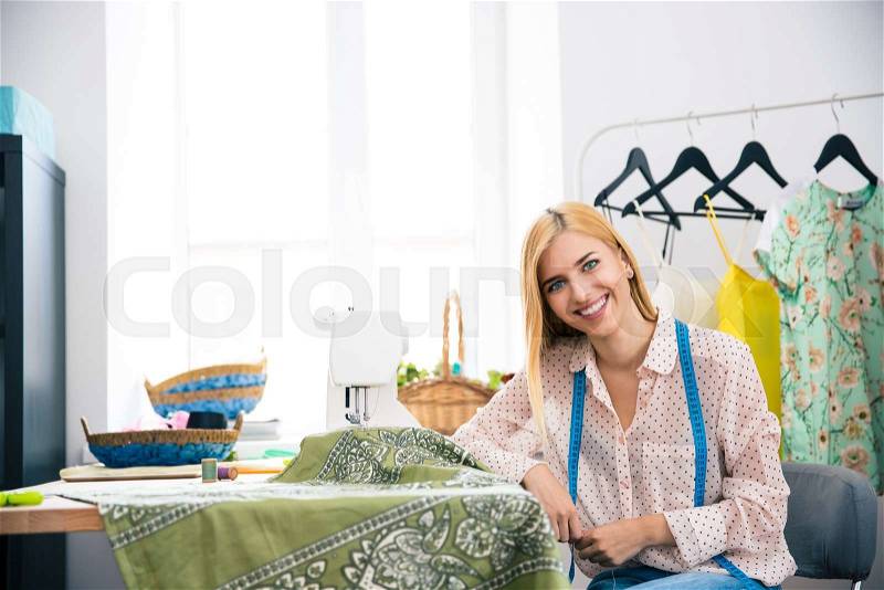 Happy female tailor sitting at her workplace in workshop and looking at camera, stock photo