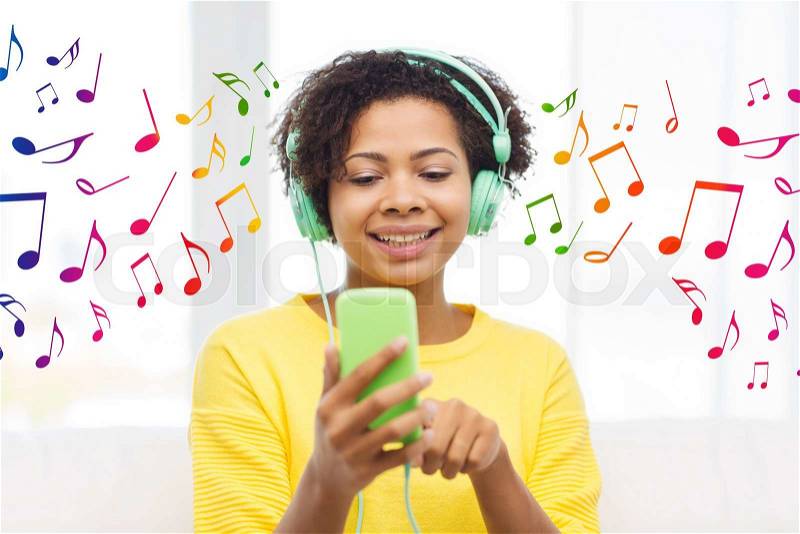People, technology and leisure concept - happy african american young woman sitting on sofa with smartphone and headphones listening to music at home over notes background, stock photo