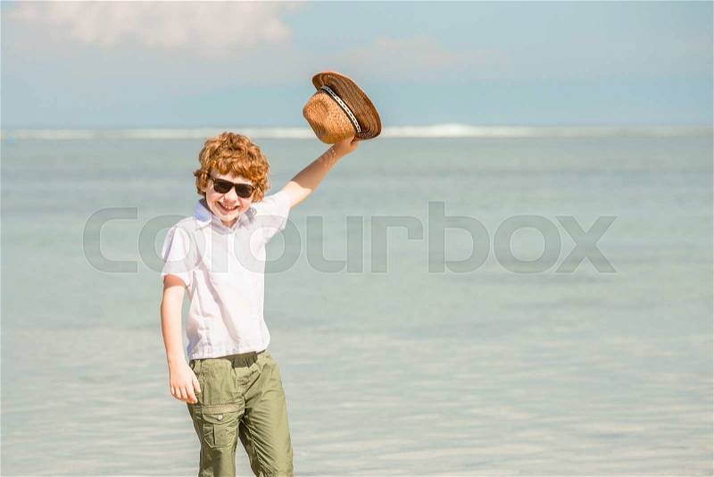 Happy red haired kid boy wearing hipster sunglasses waving hat enjoying beautiful summer sunny day stays on the beach in shallow water. Happiness, travel, vacation concept, stock photo