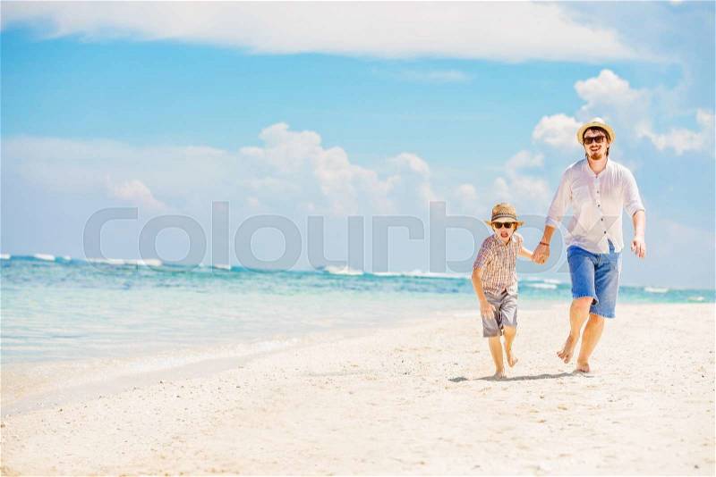 Father and son in summer nats and sunglasses running barefoot on the sea surf line having great holidays time on beautiful sunny day. Bali, Pandawa beach , stock photo