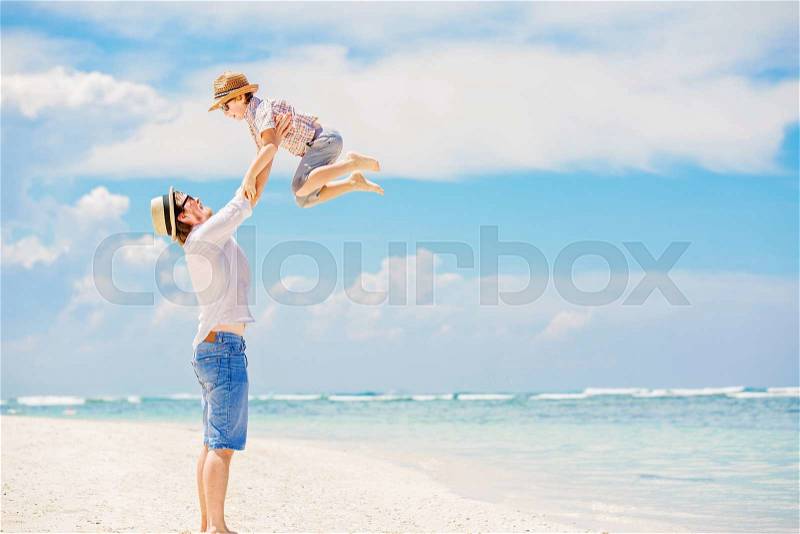 Young happy father playing with his little son standing barefoot at the beach with ocean and beautiful clouds on background. Having fun with the kid in summer coast on holidays, stock photo
