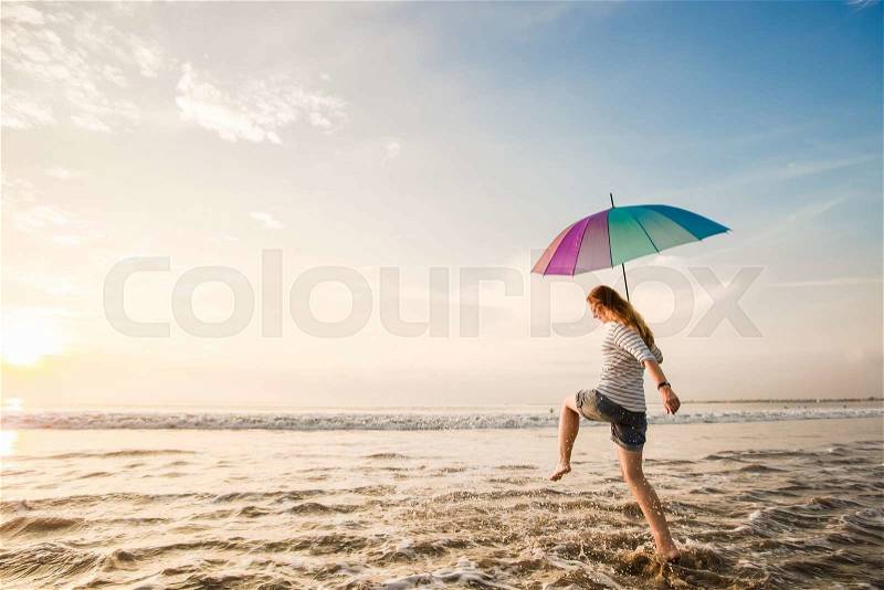 Cheerful caucasian young woman with rainbow umbrella having fun on the Jimbaran beach on Bali before sunset with beautiful ocean and blue sky on background. Travel, holidays, vacation, healthy lifestyle concept, stock photo