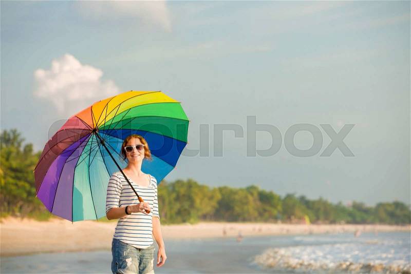 Young caucasian woman wearing white sunglasses with colourful rainbow umbrella looking at ocean on the Jimbaran beach on Bali before sunset. Travel, holidays, vacation, healthy lifestyle, tranquility concept, stock photo