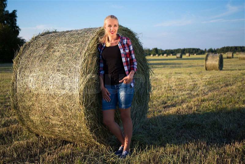 Attractive young woman relaxing outdoors standing against hay roll, stock photo