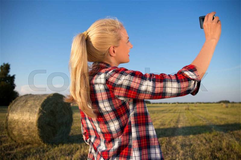 Back view of beautiful country woman making selfie photo on smartphone in field with haystacks, stock photo