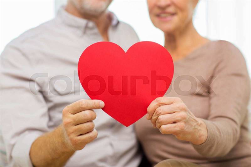 Family, holidays, valentines day, age and people concept - close up of happy senior couple holding big red paper heart shape cutout at home, stock photo