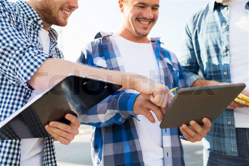 Building, construction, teamwork, technology and people concept - group of smiling builders in hardhats with tablet pc computer and clipboard outdoors, stock photo