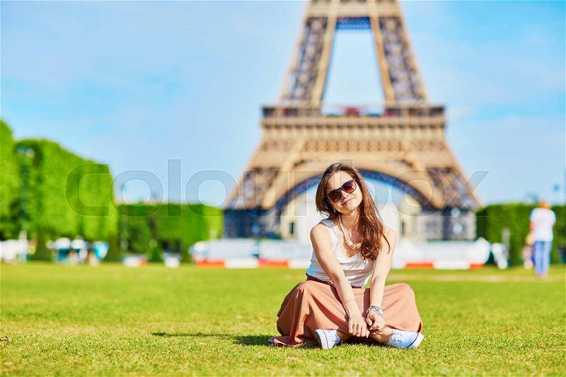 Beautiful young tourist or student girl in Paris sitting on the grass near the Eiffel tower on a summer day, stock photo